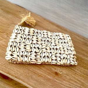 May include: A rectangular clutch covered in small, white seashells. The shells are tightly packed together, creating a textured surface. A small tassel made of natural fibers hangs from one corner. The clutch is on a wooden surface.