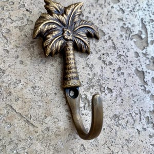 May include: A bronze-colored metal hook shaped like a palm tree. The hook features a detailed palm frond design at the top and a curved hook at the bottom. The hook is mounted on a textured, light-colored surface.