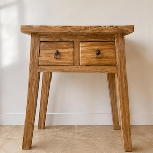 Peut inclure: Une table d'appoint en bois brun clair dotée d'un plateau rectangulaire et de quatre pieds droits. La façade de la table comprend deux tiroirs, chacun équipé d'une petite poignée en métal en forme d'anneau centrée sur la façade en bois.