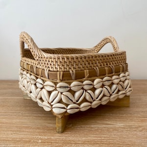 Op de afbeelding: Een rechthoekige geweven mand met twee handvatten, versierd met een rand van witte schelpen. De mand is gemaakt van lichtbruin rotan en staat op een houten voet, wat een natuurlijke, kustachtige uitstraling geeft.