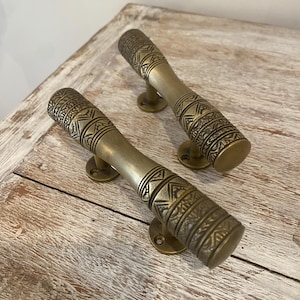 May include: Two antique brass door handles with intricate geometric patterns. The handles are cylindrical with decorative detailing and small circular mounting plates. The finish is a warm, aged brass color, suggesting a vintage or rustic aesthetic.