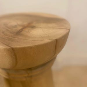 May include: A round wooden stool featuring a light tan natural wood grain surface with visible rings and a smooth, polished texture. The stool has a cylindrical base that tapers slightly, showcasing the organic patterns of the wood material.