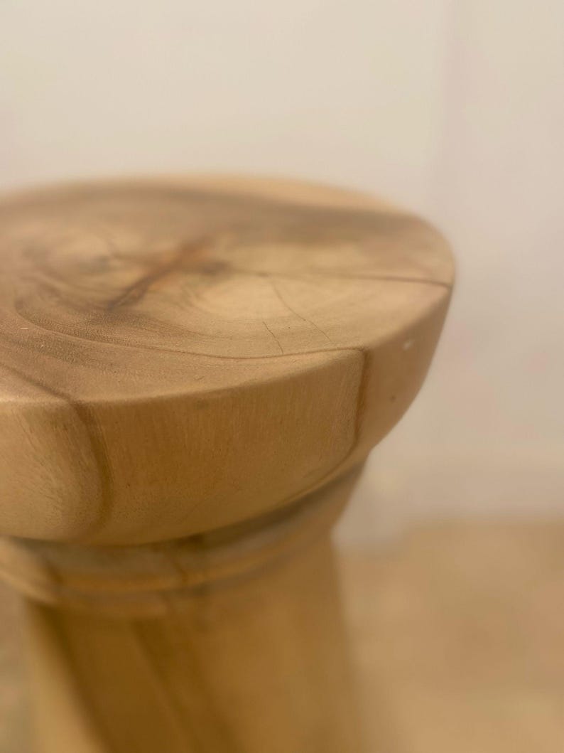 May include: A round wooden stool featuring a light tan natural wood grain surface with visible rings and a smooth, polished texture. The stool has a cylindrical base that tapers slightly, showcasing the organic patterns of the wood material.