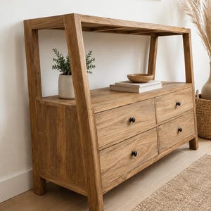 Peut inclure: Une console en bois brun clair dotée d'une étagère supérieure ouverte et de quatre tiroirs avec de petits boutons ronds noirs. La structure présente des supports latéraux inclinés et le grain du bois est visible sur tout le meuble.