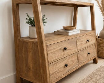 Peut inclure: Une console en bois brun clair dotée d'une étagère supérieure ouverte et de quatre tiroirs avec de petits boutons ronds noirs. La structure présente des supports latéraux inclinés et le grain du bois est visible sur tout le meuble.