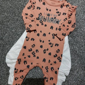 Könnte beinhalten: Ein pfirsichfarbener Baby-Strampler mit schwarzen Leopardenflecken und dem Text "I love my Mummy" in silbernem Glitzer. Der Strampler hat lange Ärmel mit Rüschen an den Schultern. Darunter ein weißer Body mit kleinen grauen Punkten.