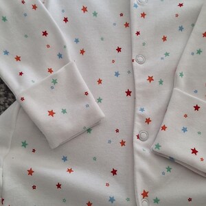 May include: White baby sleepsuit with a red trim. It features a pattern of small red, blue, and green stars. The sleepsuit has long sleeves and fastens with poppers down the front. The measurements are in cm.