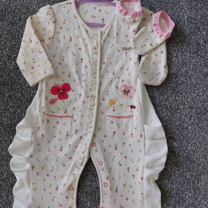 Könnte beinhalten: Cremefarbener Baby-Strampler mit floralem Muster und rosa Akzenten. Der Strampler hat gestickte Blumen, Rüschenkanten und eine Knopfleiste. Er hängt an einem lila Kleiderbügel.