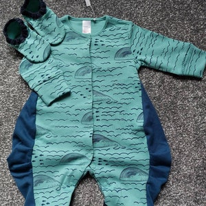 Könnte beinhalten: Ein tealfarbener Baby-Schlafanzug und passende Booties mit einem Meeresmotiv. Der Schlafanzug hat eine Knopfleiste vorne und lange Ärmel. Das Design umfasst Wale, Regenbogen und Fische. Die Booties haben eine dunkelblaue Einfassung.