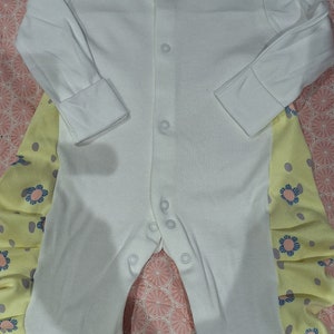 Könnte beinhalten: Weißer Baby-Strampler mit langen Ärmeln und Druckknöpfen vorne. Die Beine haben gelbe und floral gemusterte Akzente. Der Strampler liegt auf einer rosa, floral gemusterten Oberfläche.