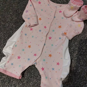 Könnte beinhalten: Rosa Baby-Schlafanzug und passende Socken mit bunten Sternen. Der Schlafanzug hat lange Ärmel und Druckknöpfe vorne. Die Socken haben rosa Bündchen und Stern-Akzente. Ein bequemes Outfit für ein Baby.