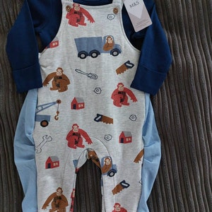 May include: A gray and blue baby outfit with a long-sleeved shirt and overalls. The overalls have a monkey and construction theme with red, blue, and yellow details. The shirt has a navy blue color. The outfit has an M&S tag.