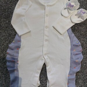 Könnte beinhalten: Ein weißer Baby-Strampler mit langen Ärmeln und Druckknöpfen. Der Strampler hat eine hellblaue Seite mit floralem Muster und Rüschenbesatz. Passende weiße Babyschuhe mit lila Schleifen sind ebenfalls enthalten.