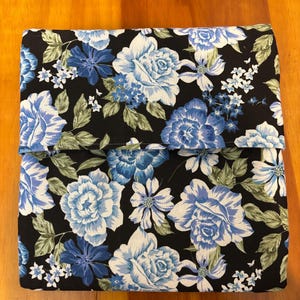 May include: A folded fabric item with a black background and a floral pattern. The pattern features large blue and white roses, smaller blue flowers, and green leaves. The fabric is folded over, creating a layered effect.