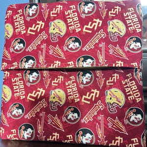 May include: A red and gold fabric item featuring repeating patterns of the Florida State Seminoles logo, helmet, and spear. The words "FLORIDA STATE" and "Seminoles" are also visible. The item appears to be a folded pouch or bag.
