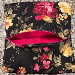 May include: A black floral clutch with a vibrant pink interior. The exterior features a pattern of red, yellow, and pink flowers with green leaves. The clutch is rectangular with a top opening.