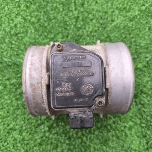 Könnte beinhalten: Ein gebrauchtes Autoteil, wahrscheinlich ein Luftmassenmesser, mit einem zylindrischen Metallkörper und einer schwarzen Kunststoffkomponente. Die schwarze Komponente trägt den Markennamen "HITACHI" und weiteren Text. Das Teil scheint gebraucht und leicht verschmutzt zu sein.