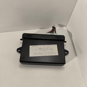 May include: A black Saab automotive electronics unit with a white label displaying text. The unit has a rectangular shape with a connector and wires extending from the top. The label includes the text "PMM UNIT 12V" and "AB AUTOMOTIVE ELECTRONICS."