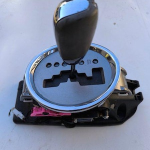May include: An automotive automatic transmission gear shifter assembly featuring a black handle with a chrome top, a circular silver gear indicator plate with P, R, N, D, 3, L markings, and a black plastic base with exposed internal components.