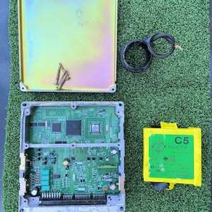 May include: An open electronic control unit (ECU) with a green circuit board, a yellow metal cover, and a yellow module labelled "C5". The ECU is on a green surface. Includes screws and wiring.