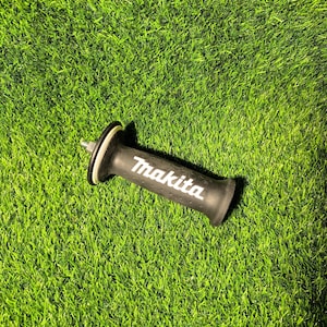 Puede incluir: Mango negro para herramienta eléctrica Makita con el logotipo blanco, sobre hierba verde. El mango tiene forma cilíndrica con un accesorio metálico y un agarre de goma. El logotipo de Makita es claramente visible.