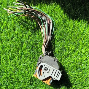 Puede incluir: Un conector eléctrico negro y gris con múltiples cables de colores que se extienden desde él. Los cables son verdes, marrones, amarillos y blancos. El conector tiene un acento naranja y está sobre hierba verde.