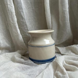 May include: A small, off-white ceramic jar with a slightly flared rim and a cylindrical body. The jar features two horizontal blue stripes, one near the top and one near the base. The jar is set against a soft, white, draped fabric background.