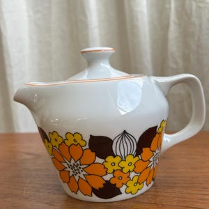 May include: White ceramic teapot with a lid and handle. The teapot is decorated with a floral pattern in shades of orange, yellow, and brown. An orange stripe accents the rim and lid. The teapot is on a wooden surface.