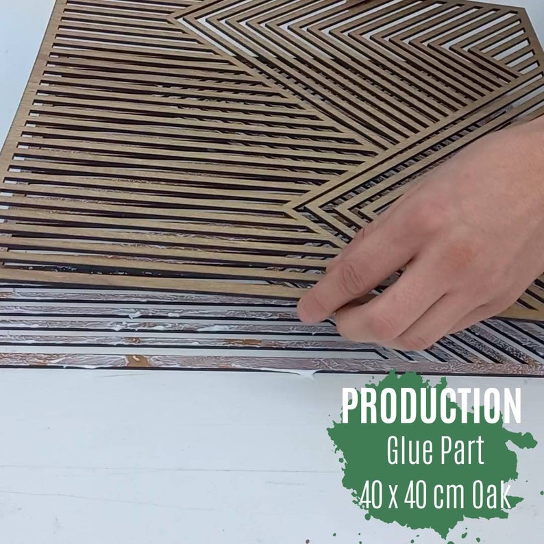 May include: A close-up of a hand applying glue to a wooden geometric wall art piece. The piece is made of oak and measures 40 x 40 cm. The text "PRODUCTION Glue Part 40 x 40 cm Oak" is visible in the image.