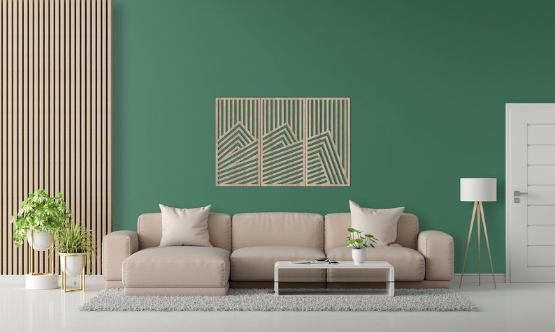 May include: A beige sectional sofa with two cushions sits in front of a green wall with a wooden wall art piece featuring a geometric mountain design. The wall art is made of three panels. A small potted plant sits on a coffee table in front of the sofa. A floor lamp with a white lampshade stands in the corner of the room.