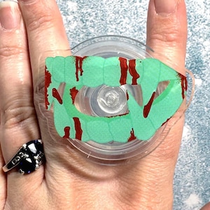 May include: A clear, circular phone grip with a green vampire teeth design and red blood accents. The phone grip is attached to a hand, and a black and silver ring is also visible.