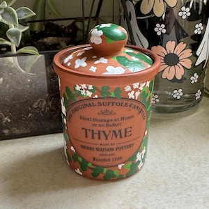May include: A brown ceramic canister with a white lid and green floral design. The canister is labeled "The Original Suffolk Canister" and "Thyme". It also says "Made by Henry Watson Pottery, England, Founded 1800".