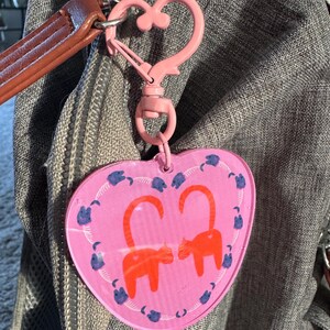May include: Pink heart-shaped keychain with a pink metal clasp. The keychain features a design of two orange cats facing each other with their tails curled up.