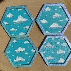 Puede incluir: Cuatro posavasos hexagonales con fondo azul y nubes pintadas de blanco. Los posavasos tienen un borde metálico plateado.