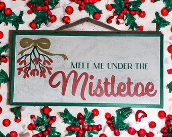 May include: A white wooden sign with green trim and a red and gold mistletoe design. The sign reads "Meet me under the mistletoe".