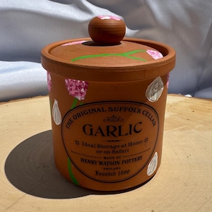 May include: Terracotta jar with a lid, hand-painted with pink flowers and white garlic cloves. The jar has a black label that reads "The Original Suffolk Cellar Garlic * Ideal Storage at Home or on Safari * Made by Henry Watson Pottery England Founded 1800".