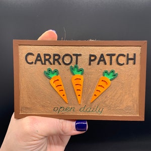 May include: A rectangular wooden sign with a brown frame and a textured, bronze-colored background. The sign reads "CARROT PATCH" in black letters, with three orange carrots and green tops. The text "open daily" is written below the carrots.