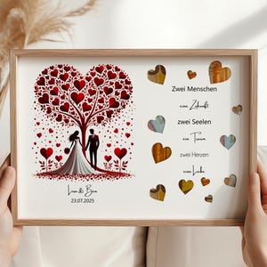 May include: A framed wedding art print featuring a tree with red heart-shaped leaves and a couple silhouette. The print includes the names "Lua & Ben" and the date "23.07.2025". Additional heart-shaped accents and text in German are also present.