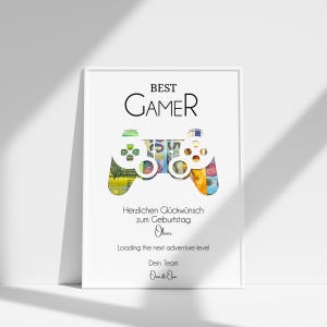 Może przedstawiać: Biały wydruk w ramie z czarno-białą ilustracją kontrolera do gier wideo wykonanego z pieniędzy. Tekst "BEST GAMER" znajduje się u g&oacute;ry wydruku. Tekst "Herzlichen Gl&uuml;ckwunsch zum Geburtstag Oliver Loading the next adventure level Dein Team Oan de O" znajduje się u dołu wydruku.