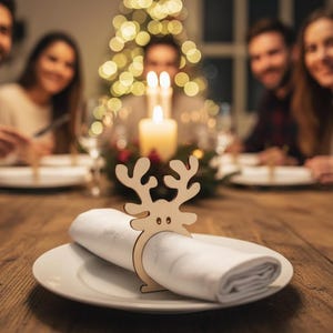May include: A wooden reindeer napkin ring holds a rolled white napkin on a white plate. The table is made of wood and is set for a holiday meal. A Christmas tree with lights and candles are in the background.