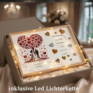 May include: A framed wedding art print featuring a couple under a heart-shaped tree, with the text "Zwei Menschen... zwei Seelen... zwei Herzen... eine Liebe." The print is illuminated by a string of LED lights and presented in a gift box.