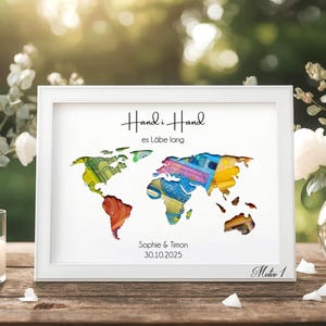 May include: A framed world map art print with a white frame. The map is a colorful collage, with text at the top and bottom. The print is displayed on a wooden table with flowers and a candle.