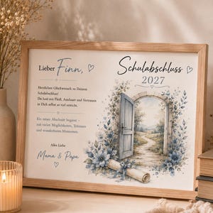 May include: A rectangular wooden frame containing a printed graduation message for Finn, dated 2027. The print features a stone archway with an open door, a winding path, blue floral accents, and a rolled diploma tied with a blue ribbon.