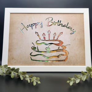 May include: A white framed wall art piece featuring a cutout design of a two-tiered cake with five lit candles and cherries on top. The text "Happy Birthday" is cut out above the cake, revealing a colorful, patterned paper background behind it.