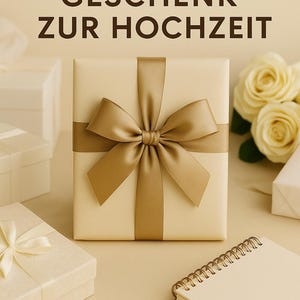 Peut inclure: Une bo&icirc;te cadeau cr&egrave;me, orn&eacute;e d'un ruban et d'un n&oelig;ud en satin dor&eacute;. Le texte "DAS PERFEKTE GESCHENK ZUR HOCHZEIT" est en haut. D'autres bo&icirc;tes cadeaux, des roses et un petit carnet sont en arri&egrave;re-plan.