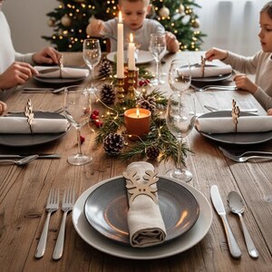 May include: A festive Christmas dinner table setting with plates, silverware, and wine glasses. Napkins are held by wooden reindeer napkin rings. A lit candle and a Christmas tree with lights are in the background. The table is made of wood.