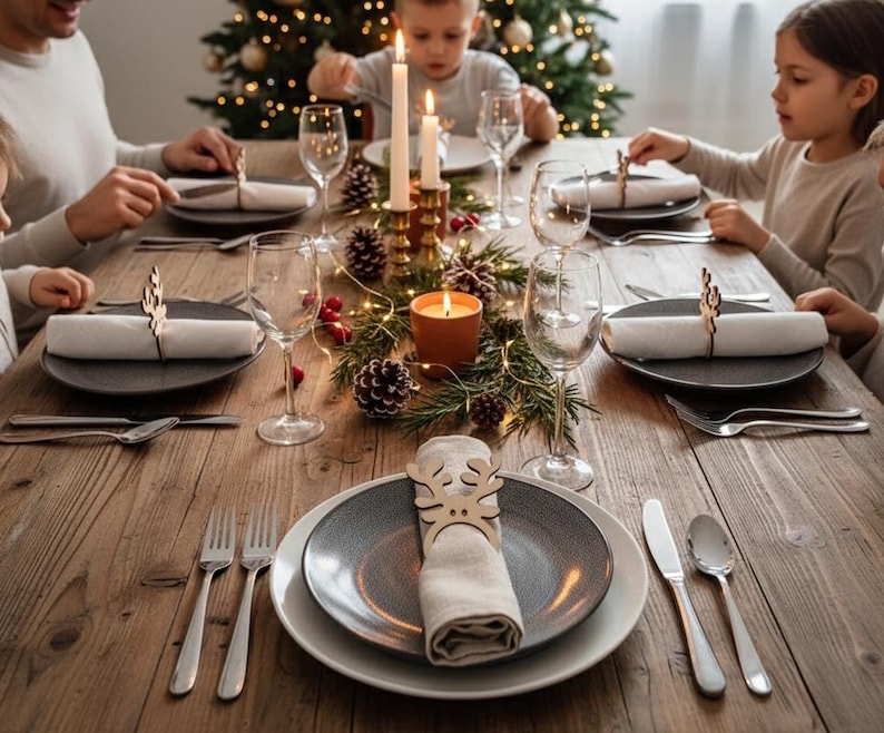 May include: A festive Christmas dinner table setting with plates, silverware, and wine glasses. Napkins are held by wooden reindeer napkin rings. A lit candle and a Christmas tree with lights are in the background. The table is made of wood.