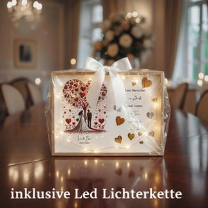 May include: A wooden framed wedding keepsake with a heart-shaped tree design, a bride and groom silhouette, and the date 23.07.2023. The frame is wrapped in clear cellophane and tied with a white ribbon. Includes LED string lights.