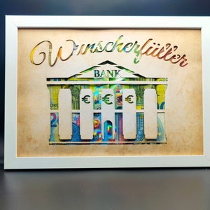 May include: A white framed picture with a brown background. The picture features a cutout of a bank building with the word "BANK" above the building. The building is made of euro bills. The text "Wunschklitter" is written in cursive at the top of the picture.