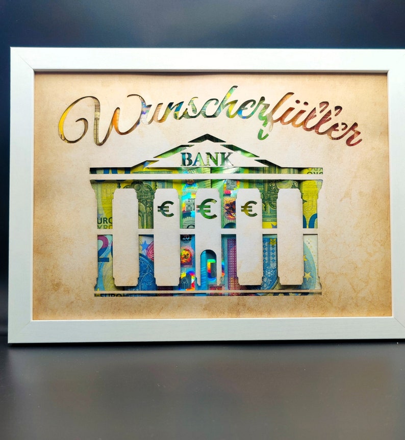 May include: A white framed picture with a brown background. The picture features a cutout of a bank building with the word "BANK" above the building. The building is made of euro bills. The text "Wunschklitter" is written in cursive at the top of the picture.